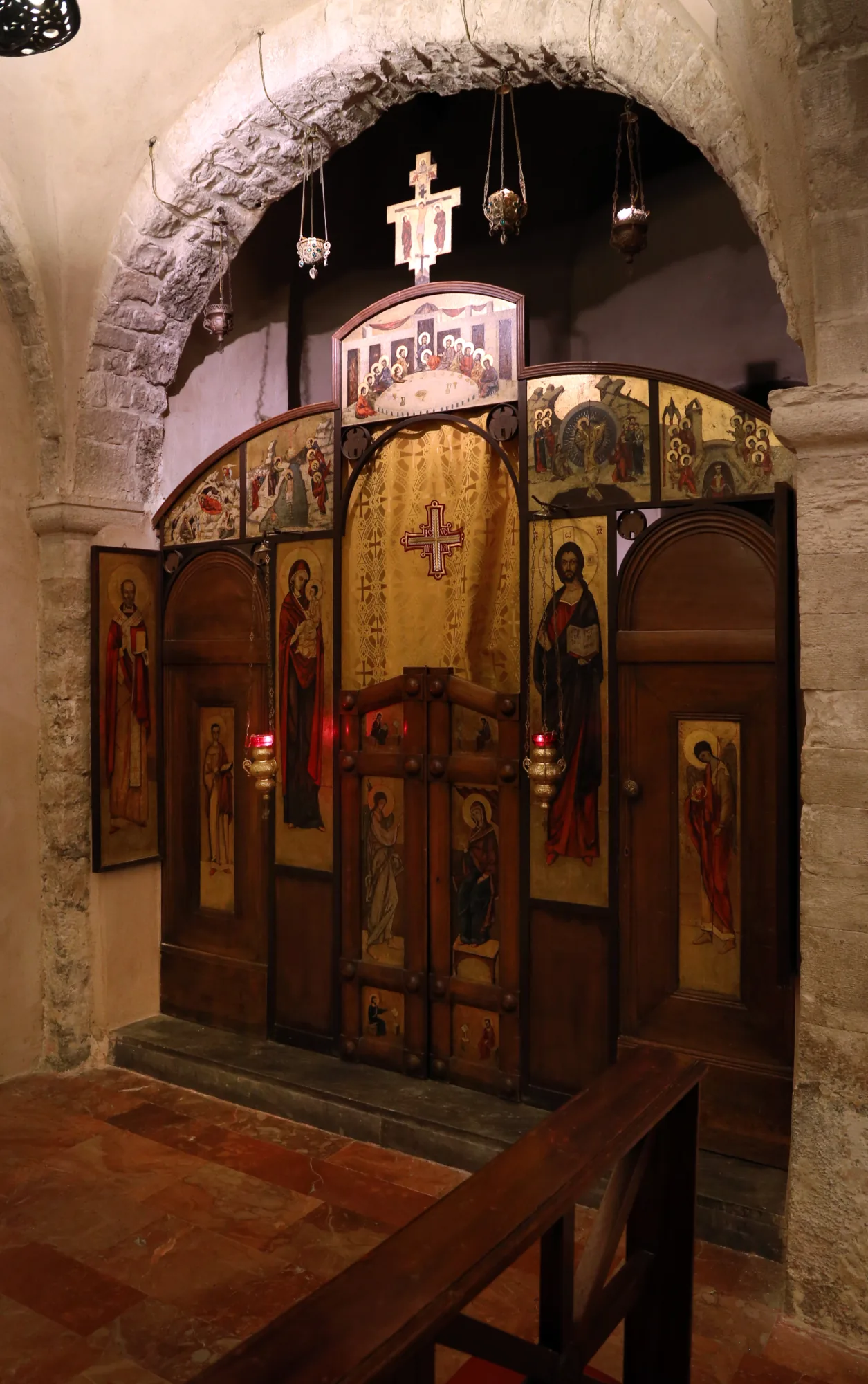 A wooden Orthodox icon screen with painted icons and central royal doors, set beneath a Romanesque stone arch in the crypt of the Basilica di San Nicola in Bari.