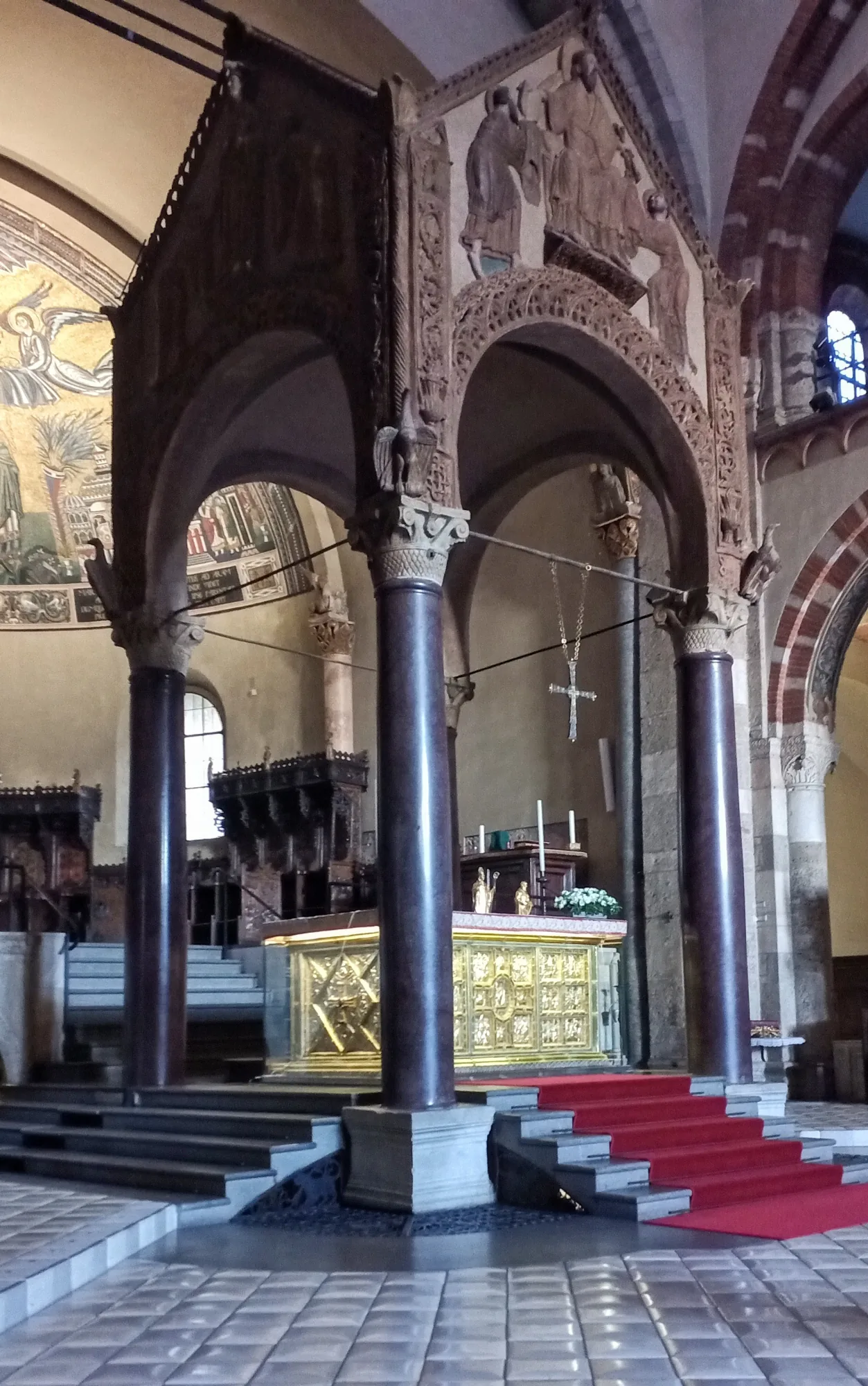 The presbytery of the Basilica di Sant'Ambrogio: a carved stone canopy on four dark porphyry columns standing over a gilded altar, with the apse mosaic visible behind it and steps leading up from the nave.