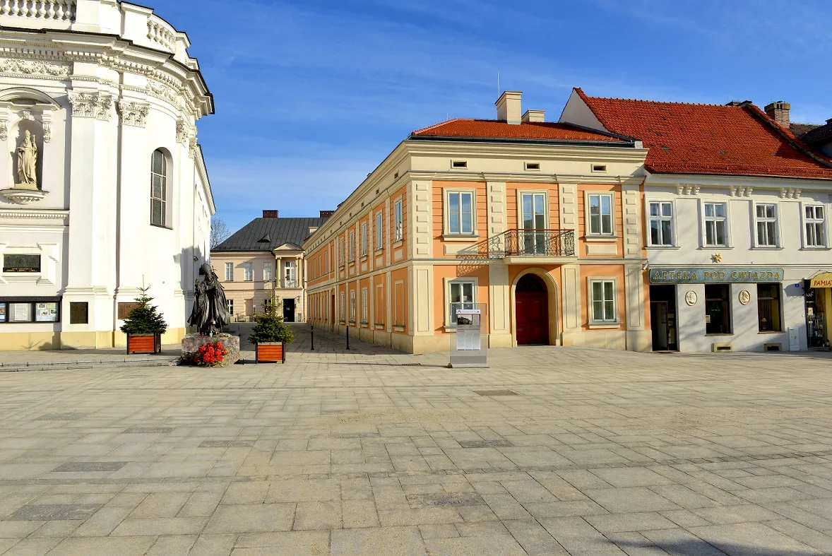 The corner of plac Jana Pawła II in Wadowice, the white flank of the basilica on the left and the ochre family-home building on the right, the square empty in low sun.