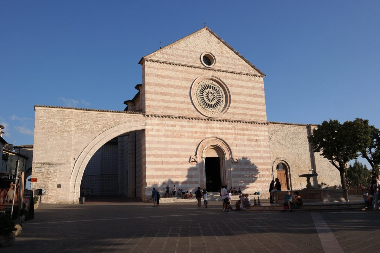 The pink and white striped facade and rose window of the Basilica of Santa Chiara in Assisi, lit by evening sun.