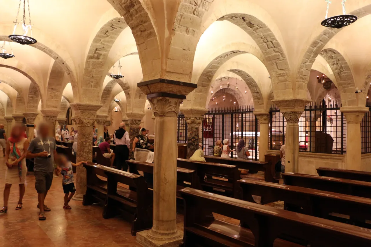 The crypt of the Basilica di San Nicola in Bari: rows of stone columns under low cross vaults, wooden benches, pilgrims standing and sitting, and an iron grille closing the tomb chapel on the right.