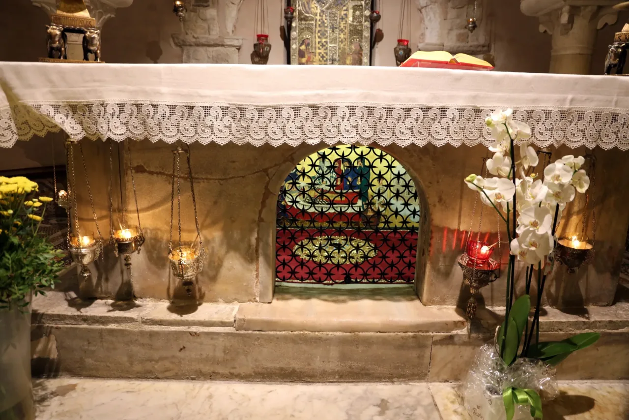 The tomb of Saint Nicholas beneath the crypt altar in Bari: a stone tomb with an arched opening in its front closed by a decorative grille, oil lamps hanging on chains to either side, flowers and a lace altar cloth above.