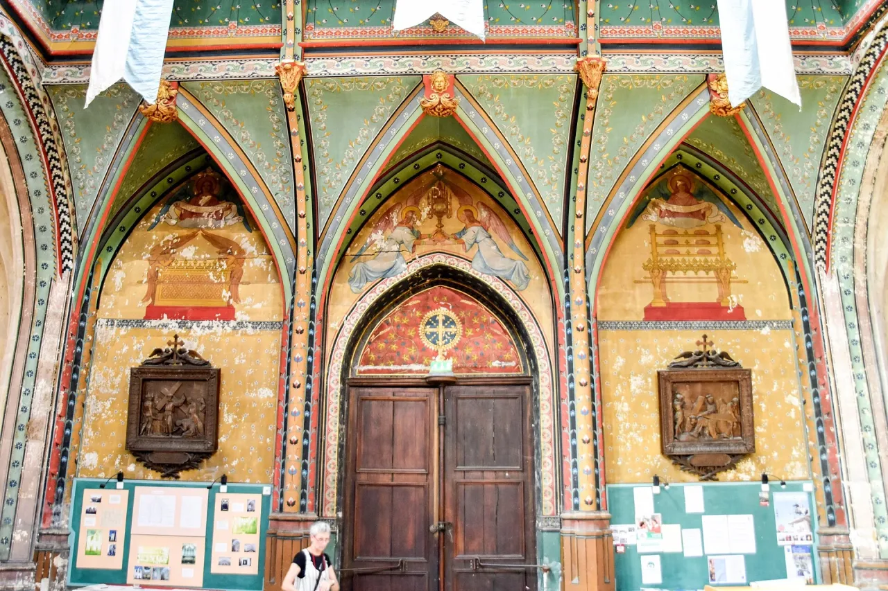 The painted west wall inside Église Saint-Aignan in Chartres, its arches and vaults covered in green, red and gold patterning above the parish notice boards.