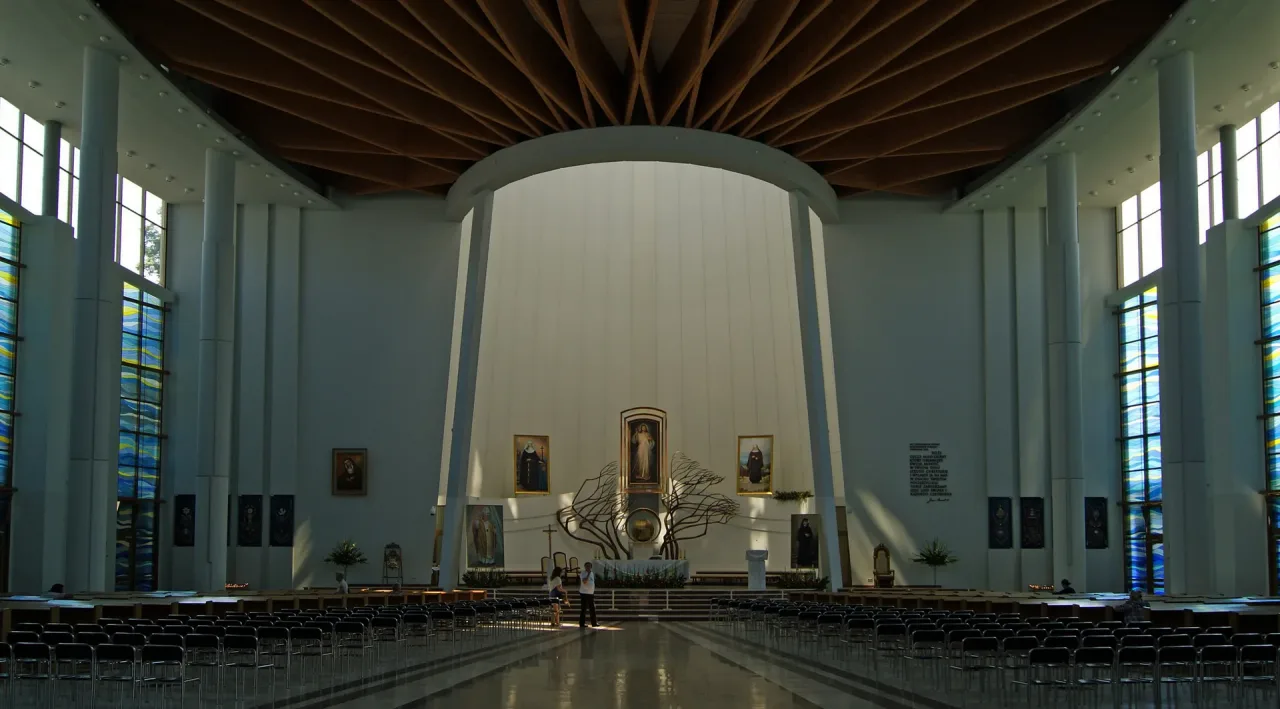 The white nave of the modern Divine Mercy basilica at Łagiewniki, its boat-shaped timber ceiling above rows of chairs facing a copy of the Merciful Jesus image.