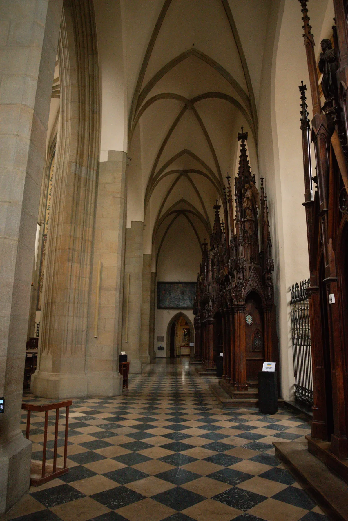 A side aisle of the Dominican basilica in Kraków, its checkered stone floor running past dark wooden confessionals toward a lit chapel.