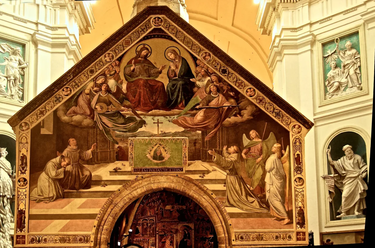 The complete Porziuncola chapel standing inside the Basilica of Santa Maria degli Angeli, with its frescoed facade and small stone body visible together.