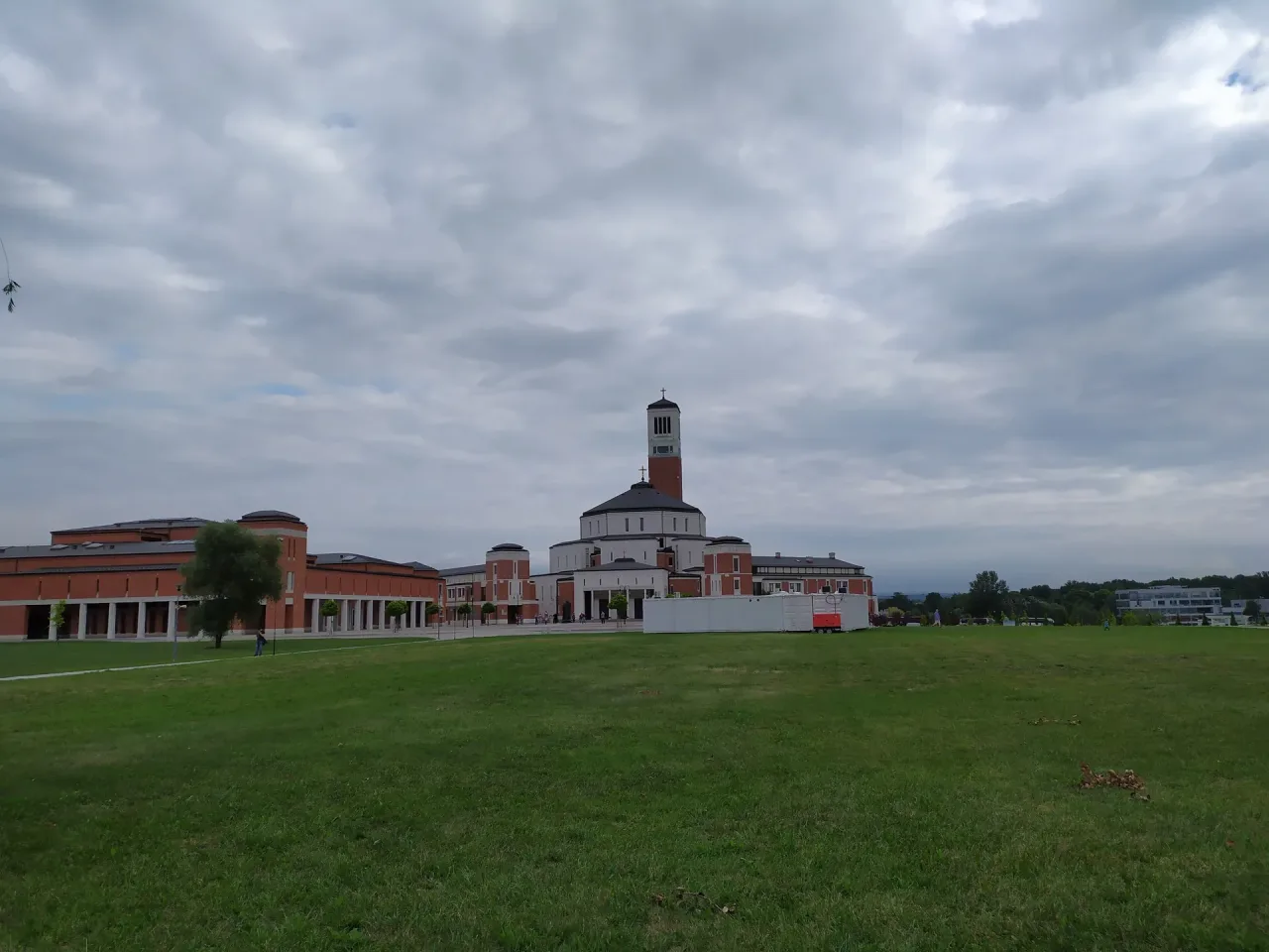 The Sanctuary of Saint John Paul II in Kraków, its dark dome and white bell tower rising above the red brick ranges of the John Paul II Centre, seen across open grass.