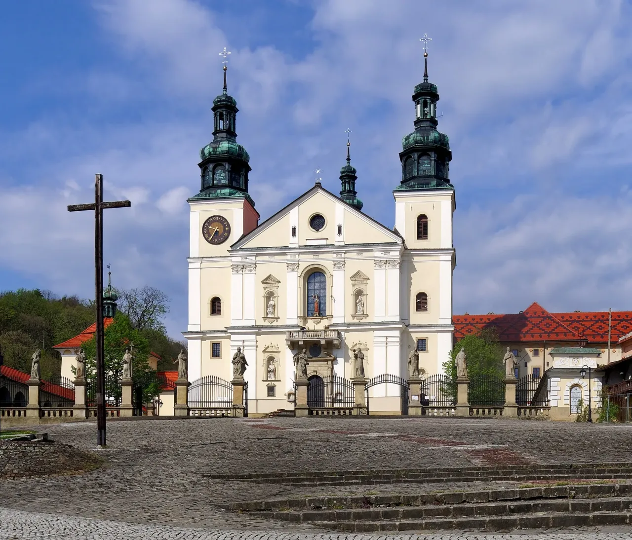 The white west front of the basilica of Our Lady of the Angels at Kalwaria Zebrzydowska, twin copper-domed towers above a pedimented façade, a tall wooden cross standing on the cobbled forecourt to the left.