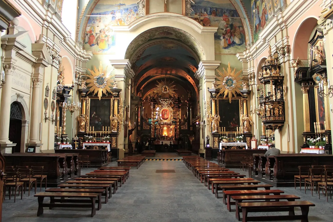 The nave of the Kalwaria basilica looking towards the high altar, painted vaults overhead, gilded side altars with sunburst frames, and plain wooden benches on a stone floor.