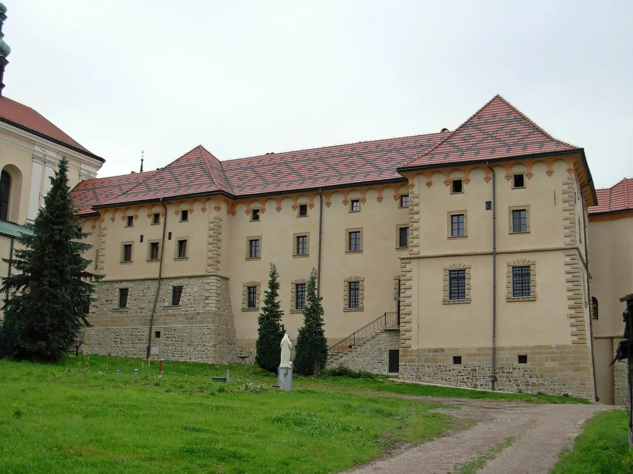 The Bernardine monastery at Kalwaria Zebrzydowska, a long cream range on a stone base with red tiled roofs and a corbelled parapet, a small white statue on the grass in front of it.