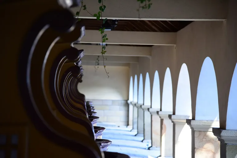 A receding row of dark carved wooden confessionals standing under the open arcade of the basilica courtyard at Kalwaria Zebrzydowska, sunlight falling through the arches onto the paving.
