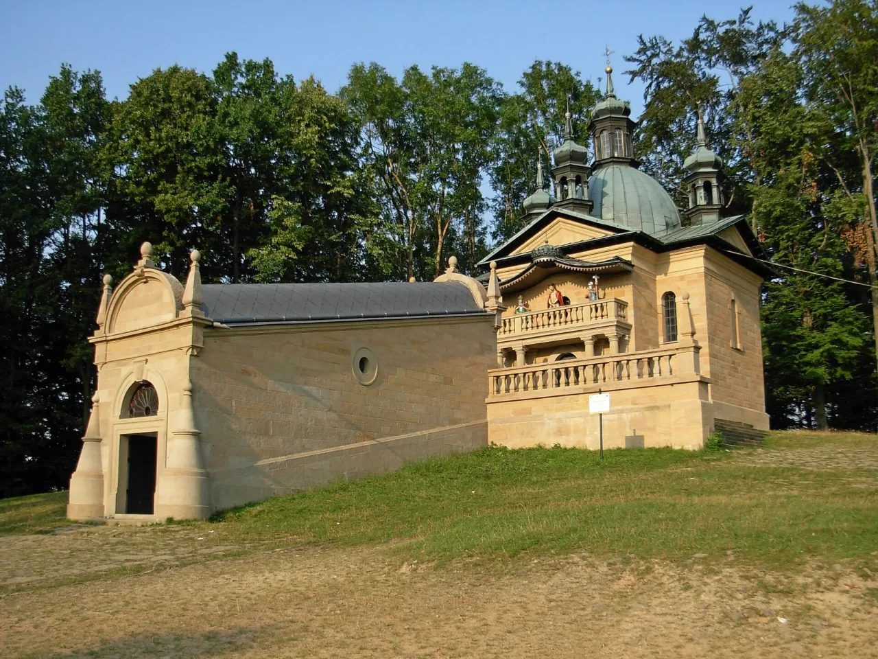 The stone chapel of Pilate's Town Hall standing on open grass among trees at Kalwaria Zebrzydowska, its domed lantern above a balustraded terrace, with the long low range of the Holy Stairs attached to the left.