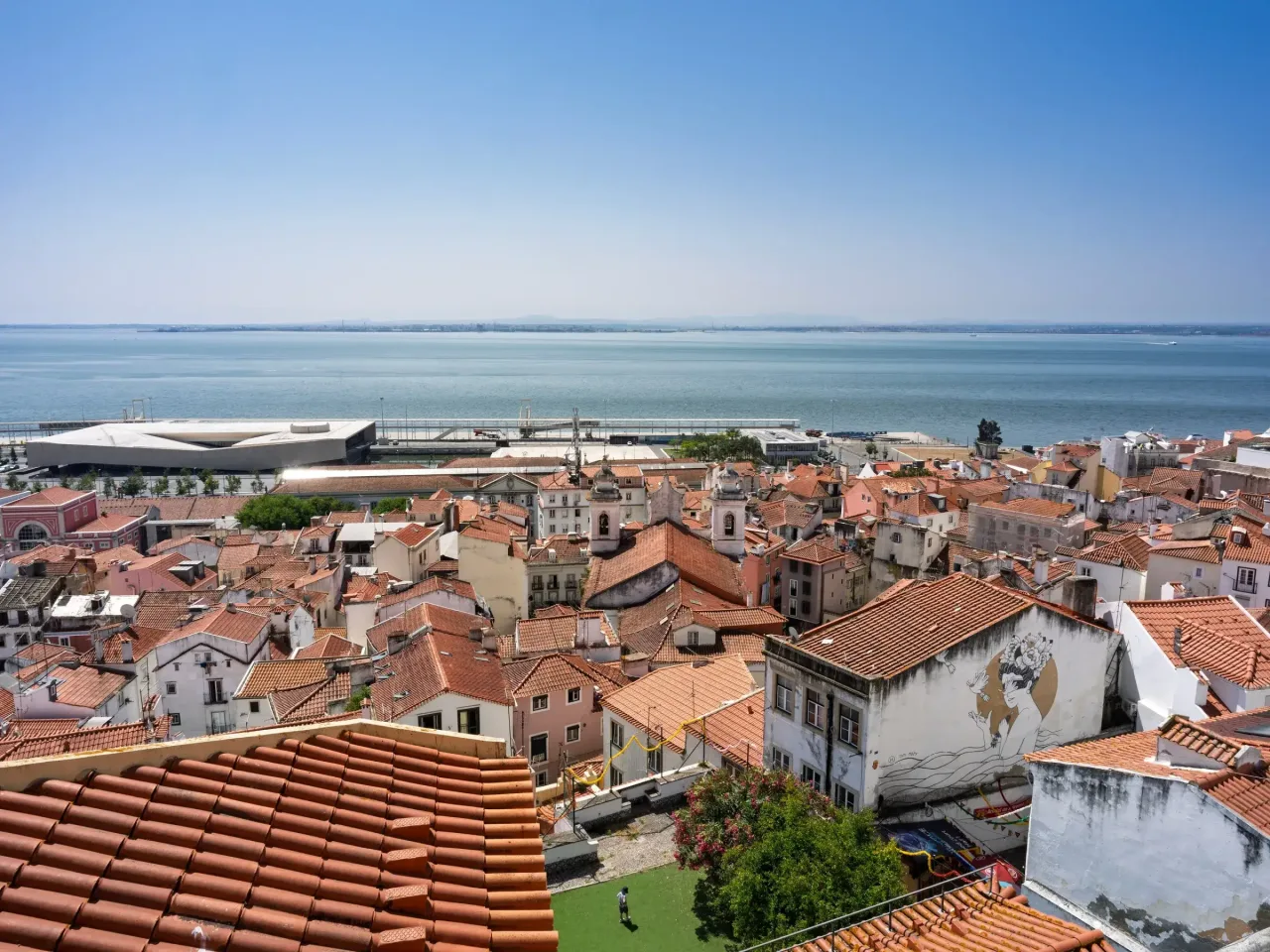 The tiled rooftops of Alfama seen from the Miradouro de Santa Luzia, with the Tagus estuary beyond.