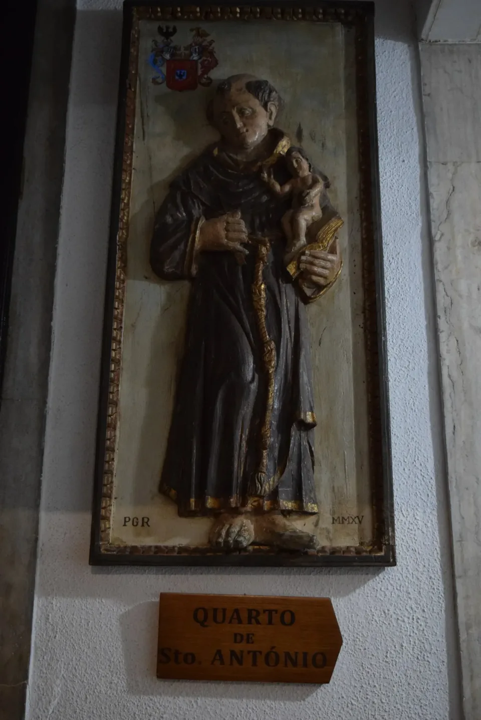 A carved and painted relief of Saint Anthony holding the Child Jesus, mounted above a wooden sign reading Quarto de Santo António.