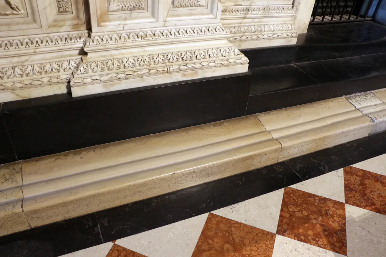 Two deep grooves worn into a pale marble step at the base of the carved enclosure, beside an inlaid marble floor.
