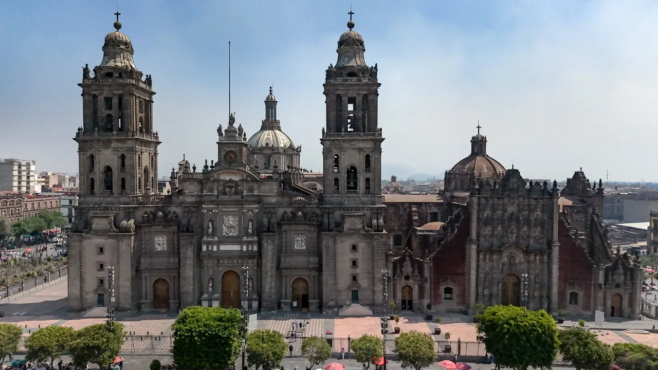 The Metropolitan Cathedral of Mexico City seen across the Zócalo, its two bell towers and central dome above a long baroque front, with the darker tezontle facade of the Sagrario Metropolitano attached at the right.