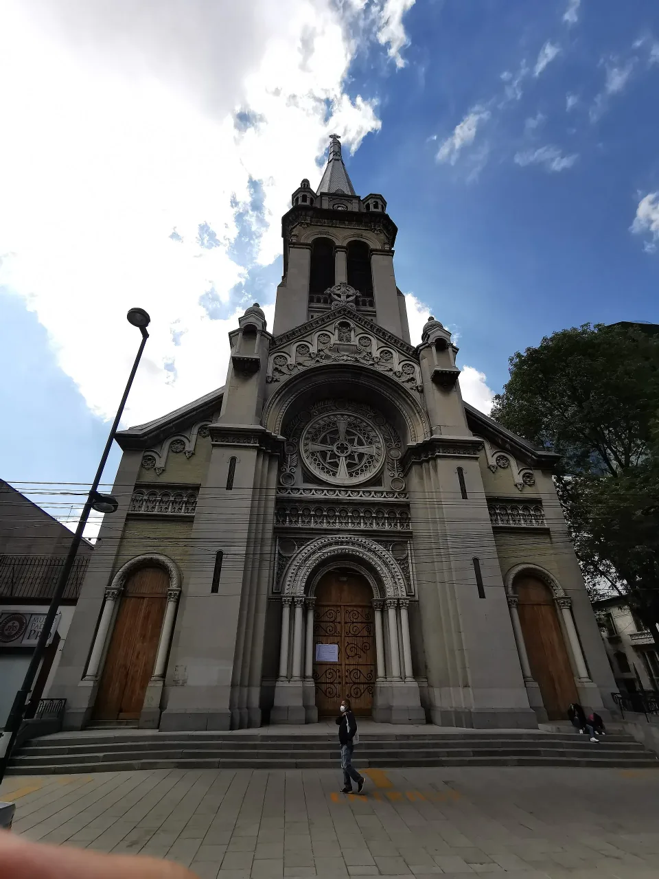 The front of the parish church of the Sagrada Familia in Colonia Roma, Mexico City, a tall early twentieth-century stone facade with a rose window, carved portal and a single spire.