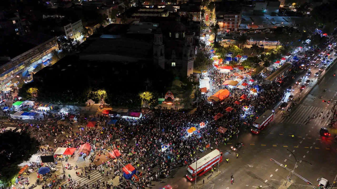 A night view from above of crowds filling the streets and plaza around the church of San Hipólito in Mexico City for the monthly devotion to Saint Jude Thaddaeus, with market stalls and buses along the avenue.
