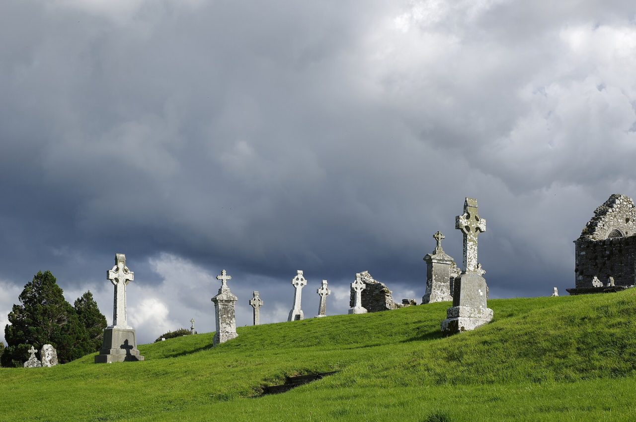 High crosses and carved grave markers standing across the open monastic field at Clonmacnoise under a darkening sky.