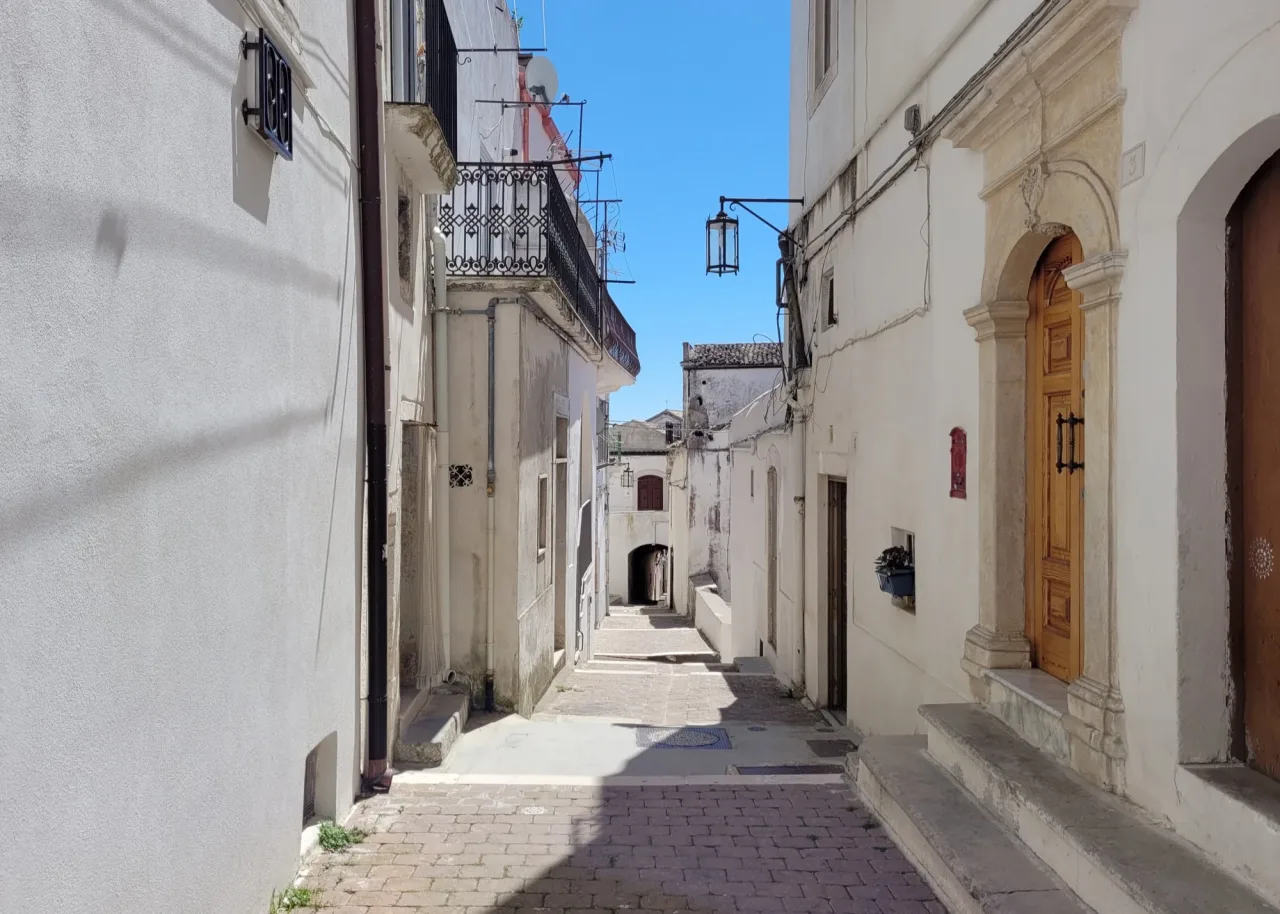 A narrow stepped street in Monte Sant'Angelo between whitewashed houses, with a stone doorway on the right, a wrought-iron lantern overhead, and an archway at the far end.