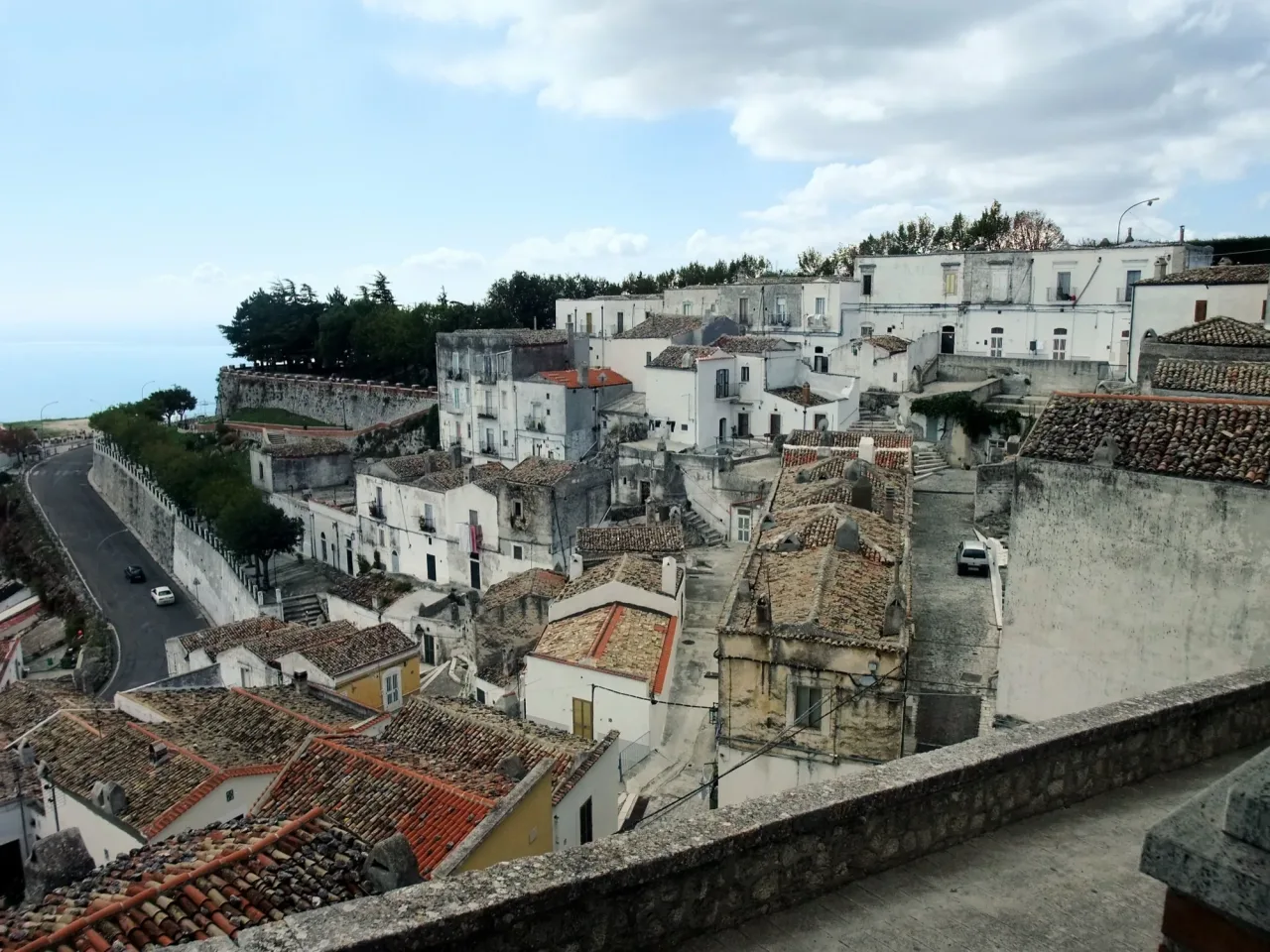 The town of Monte Sant'Angelo seen from above, white houses and stone-tiled roofs stepping down a steep slope, with a road curving along a wall and the sea flat on the horizon.