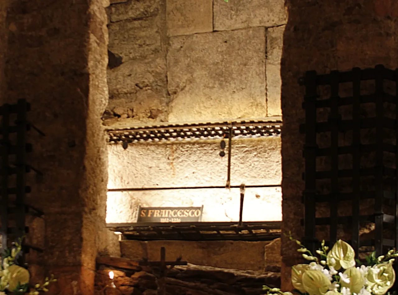 The stone tomb of Saint Francis in the crypt of the Basilica di San Francesco at Assisi, lit behind an iron grille and marked S. FRANCESCO.