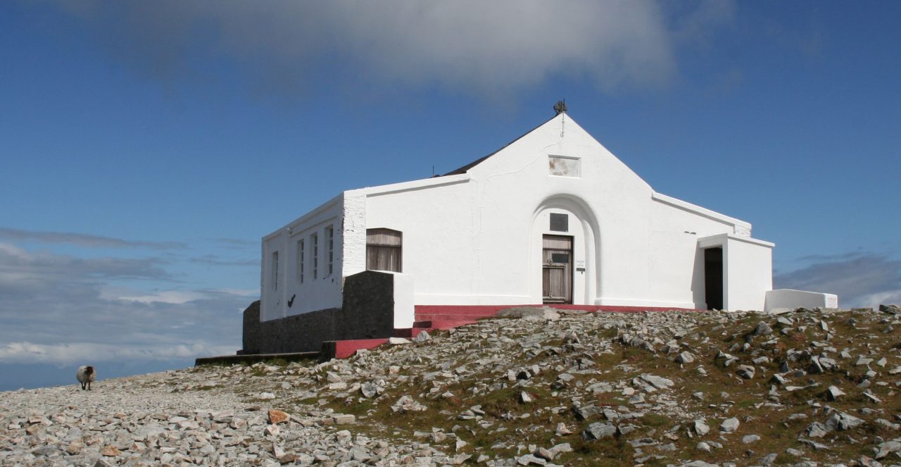 The white Saint Patrick's Oratory on the rocky summit of Croagh Patrick beneath a blue sky.