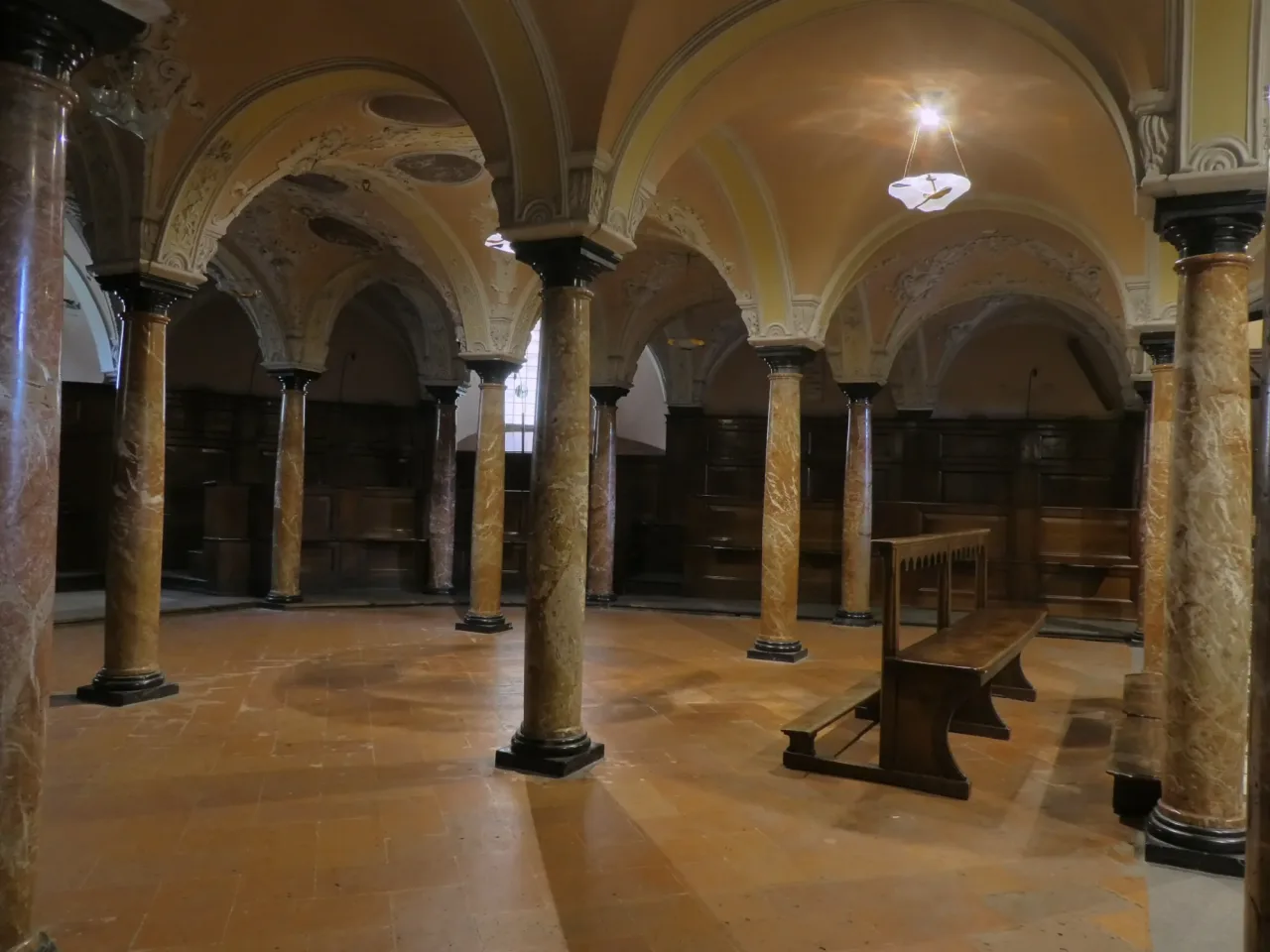 The crypt of the Basilica di Sant'Ambrogio: a low vaulted room on veined marble columns with dark wooden benches around the walls and a tiled floor.
