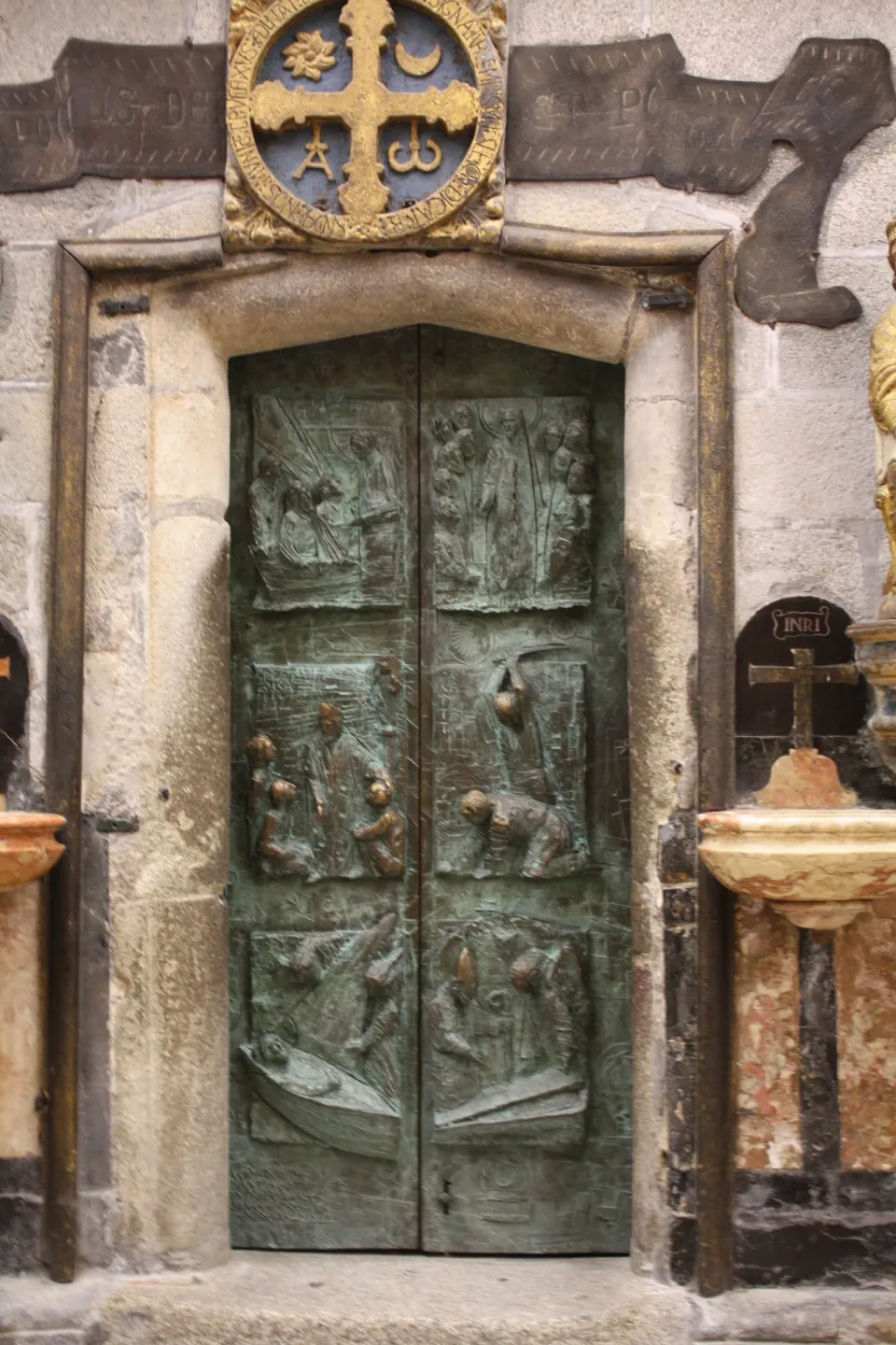 The bronze leaves of the Holy Door of Santiago Cathedral, cast in relief panels, set in a stone frame beneath a gilded cross.