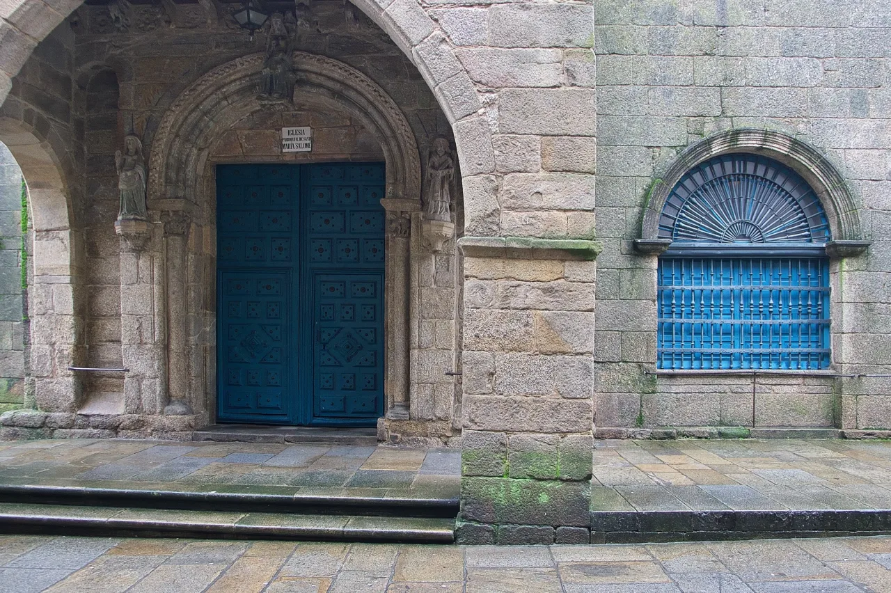 The open stone portico of Santa María Salomé in Santiago de Compostela, its blue-painted Romanesque doorway set back under a wide arch with carved figures on the jambs.