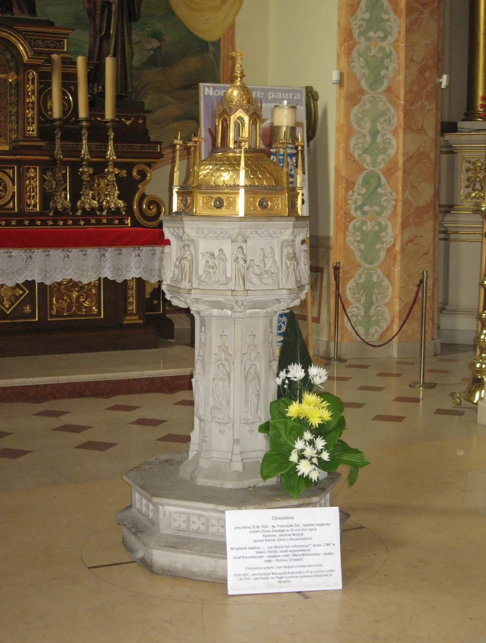 The stone baptismal font of the Wadowice basilica, carved with figures under a gilt domed cover, standing on a low step with flowers and an explanatory placard beside it.