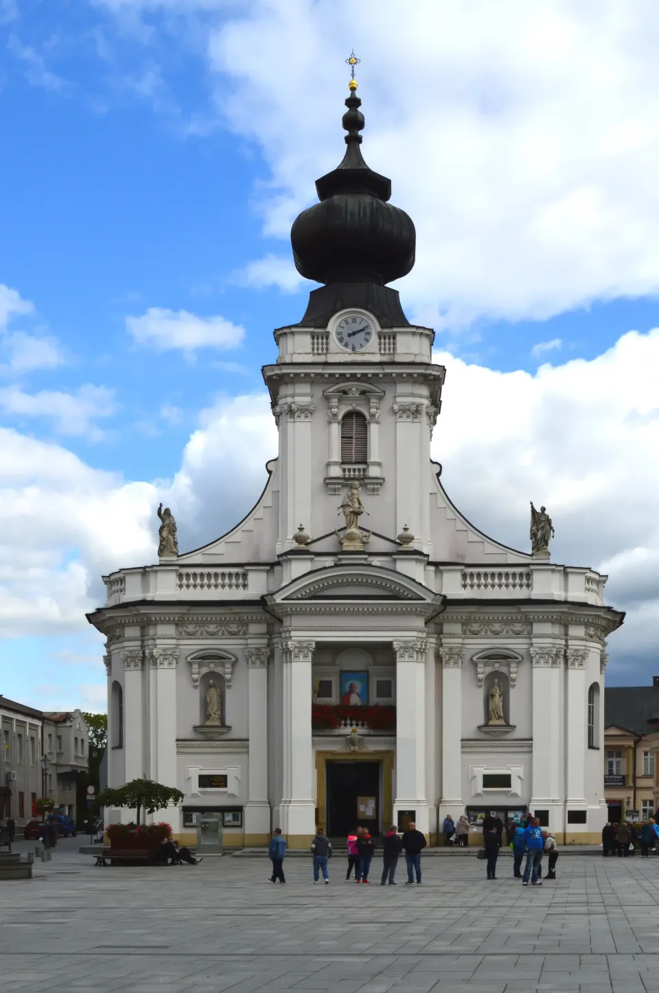 The west front of the Wadowice basilica, a white Baroque facade with paired columns, saints in niches, and a dark onion-domed tower above the clock.