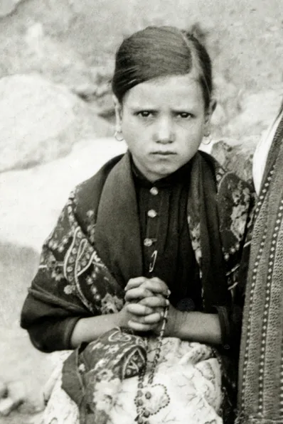 Photograph of Jacinta Marto as a young girl in a patterned shawl and headscarf, hands clasped around a rosary.